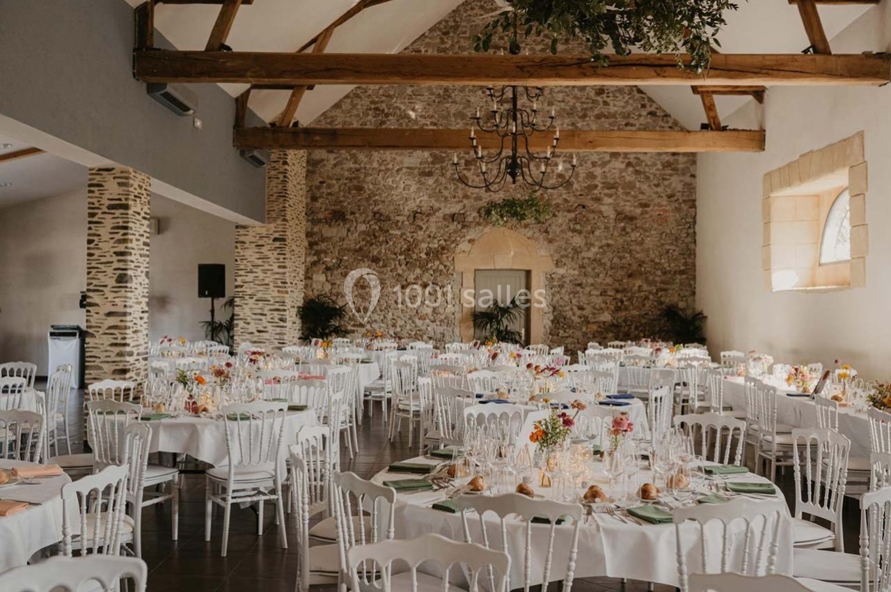 Salle de réception lumineuse avec des tables rondes décorées, des chaises blanches et des poutres apparentes au plafond.