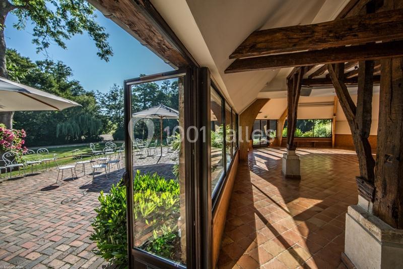 Location salle Saint-Léger-en-Bray (Oise) - Domaine du Colombier #7 Vue depuis une salle avec poutres apparentes donnant sur une terrasse ensoleillée avec tables, chaises et jardin verdoyant.