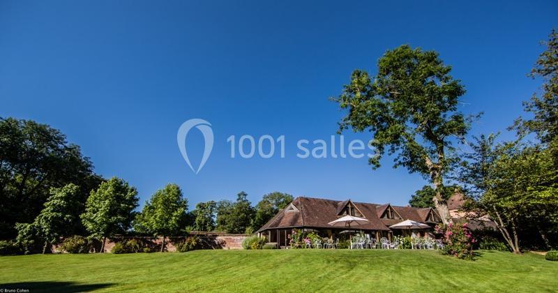 Location salle Saint-Léger-en-Bray (Oise) - Domaine du Colombier #5 Pelouse verdoyante avec une maison en briques et toit en tuiles, entourée d'arbres et de parasols sur une terrasse.