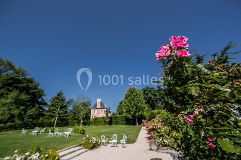 Location salle Saint-Léger-en-Bray (Oise) - Domaine du Colombier #9 Allée bordée de rosiers roses menant à un jardin avec des chaises blanches et une maison en arrière-plan.