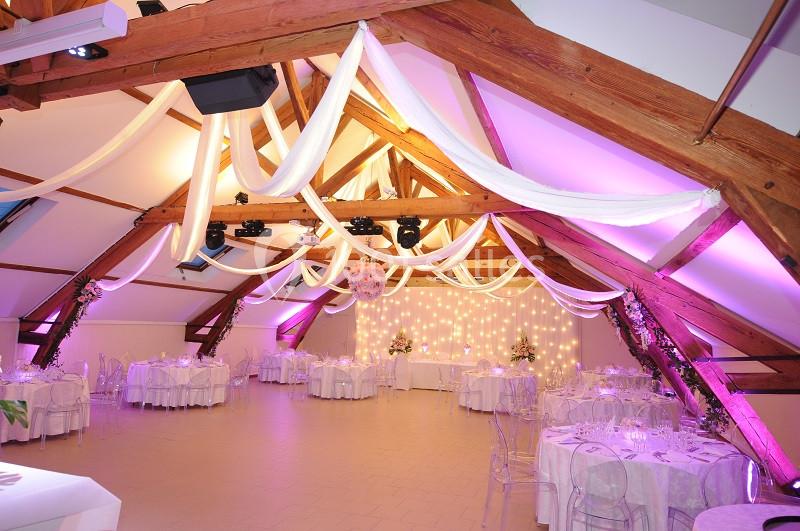 Salle de réception décorée avec des poutres apparentes, des guirlandes lumineuses et des tables dressées.