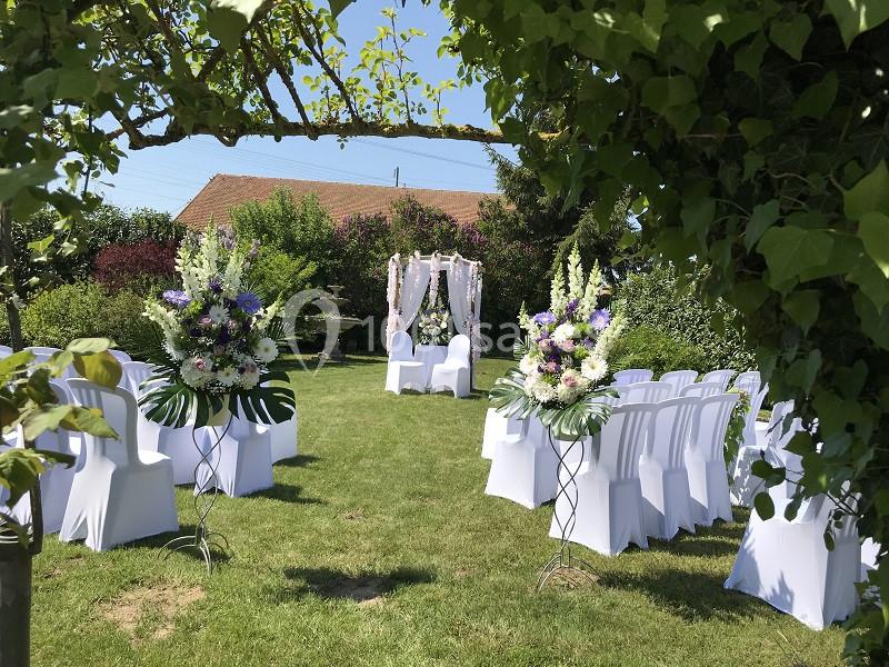 Chaises blanches alignées et arche décorée de fleurs dans un jardin verdoyant pour une cérémonie en plein air.