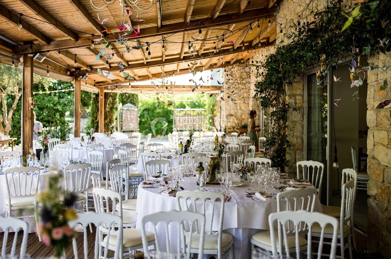 Salle de réception en plein air avec tables rondes dressées, chaises blanches et décoration florale naturelle.