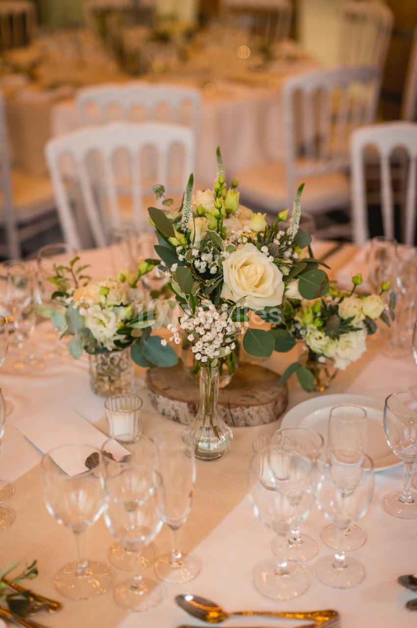 Centre de table floral avec roses blanches, gypsophile et feuillage, entouré de verres et couverts sur une nappe beige.