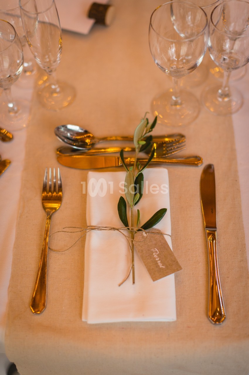 Serviette blanche pliée avec un brin d'olivier et une étiquette, entourée de couverts et verres sur une table dressée.