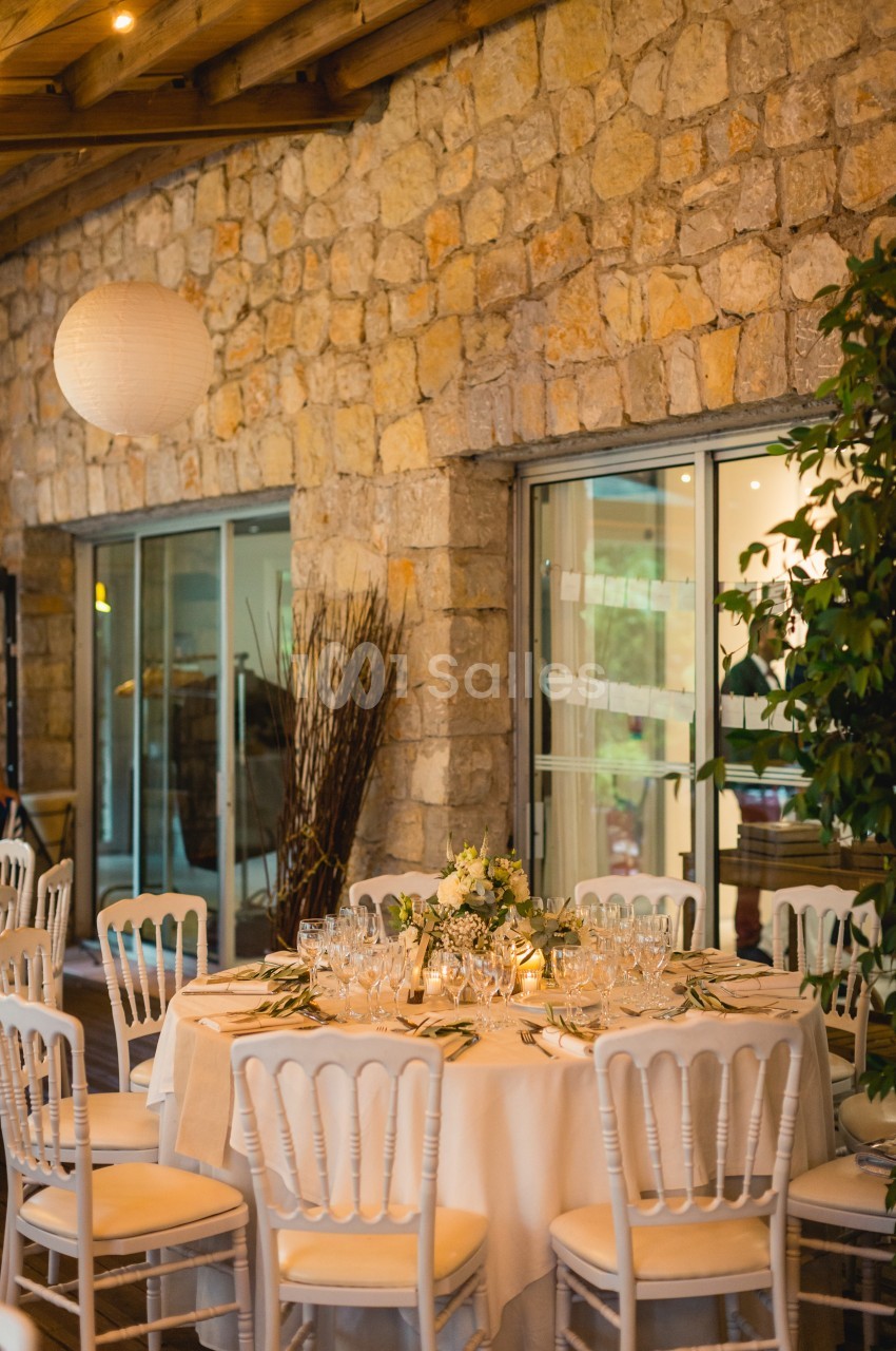 Table ronde élégamment dressée avec vaisselle raffinée, décorée de fleurs, dans une salle en pierre avec grandes baies…
