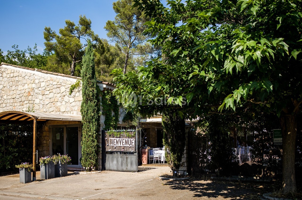 Façade en pierre d'un bâtiment entouré d'arbres, avec une pancarte ’Bienvenue’ et une terrasse ombragée.