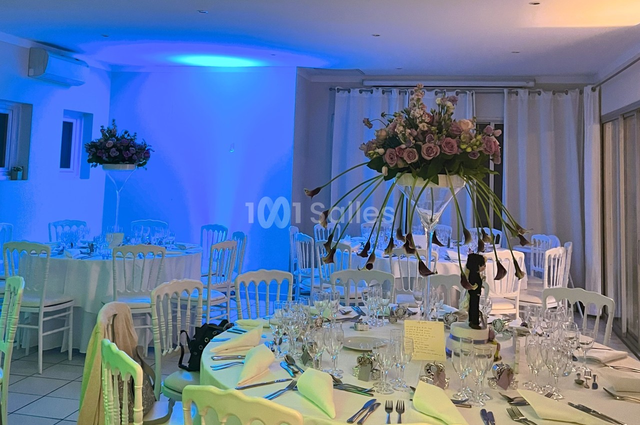 Salle de réception décorée avec des tables dressées, des chaises blanches et des centres de table fleuris.