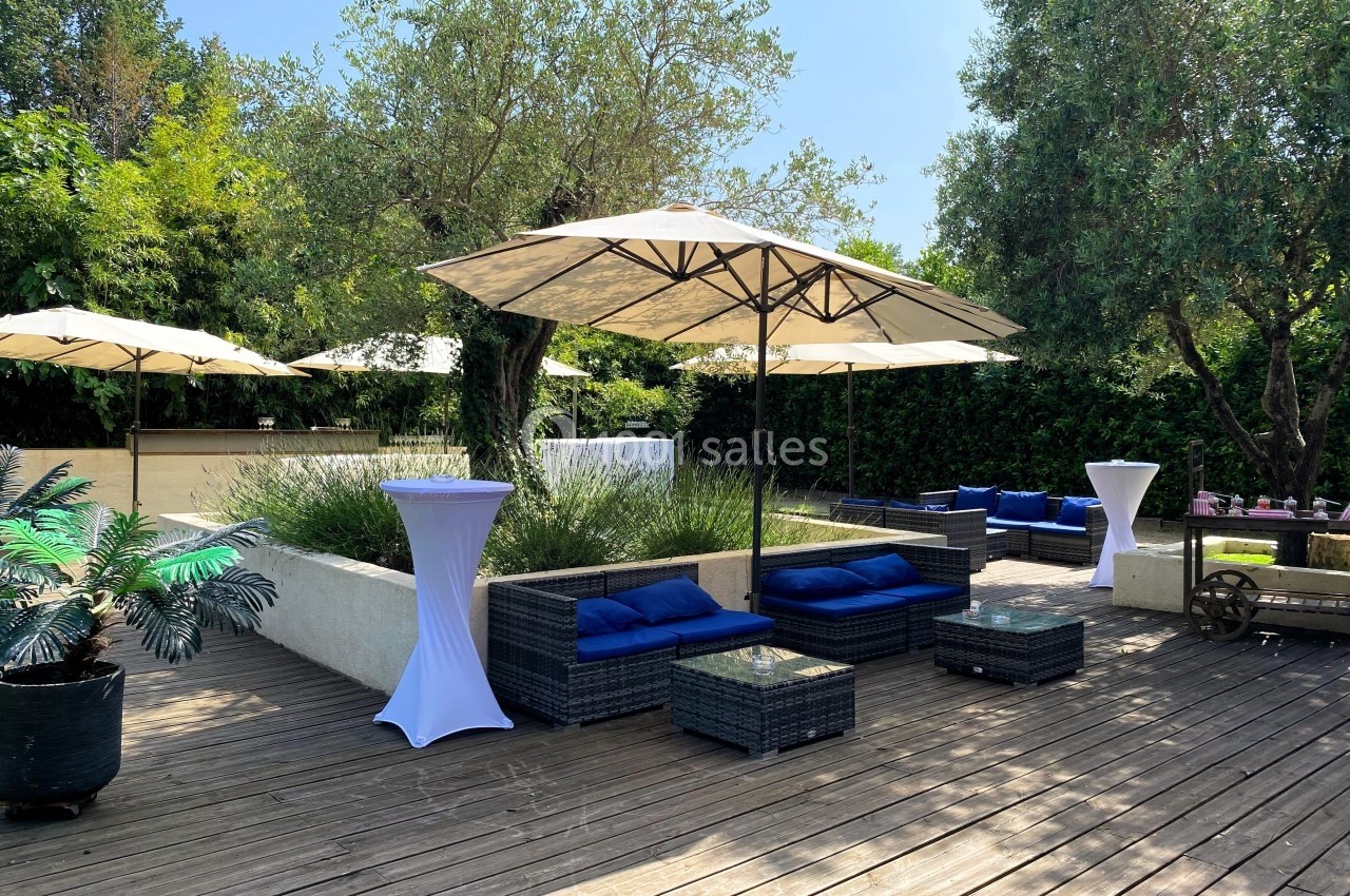 Terrasse en bois avec salons de jardin, parasols, tables hautes et végétation environnante sous un ciel dégagé.