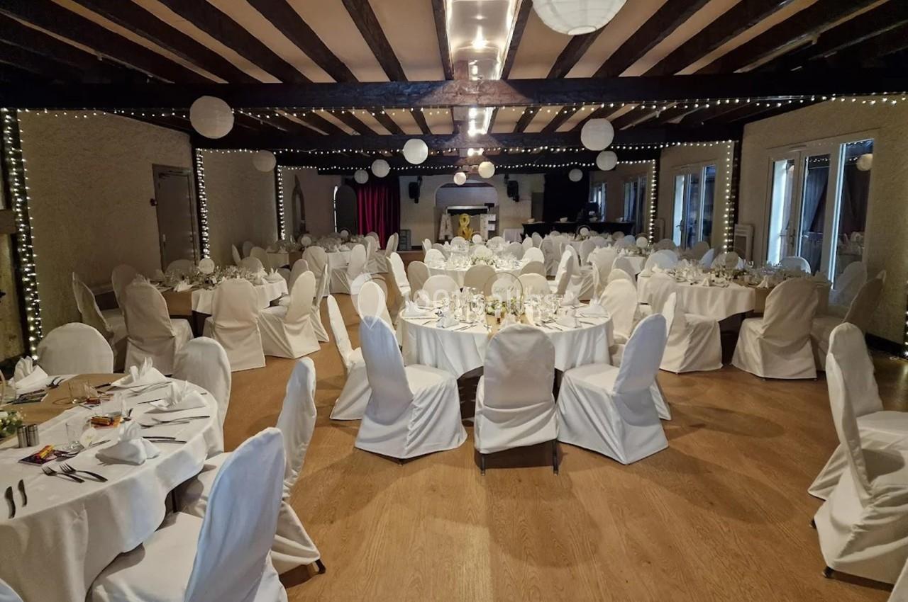 Salle de réception décorée avec des tables rondes dressées et des chaises blanches sous un plafond à poutres apparentes.