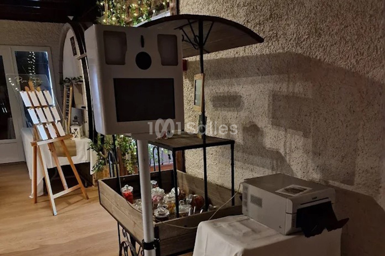 Borne photo avec écran et objectif, installée près d'une imprimante et d'un chariot décoratif dans un espace intérieur.