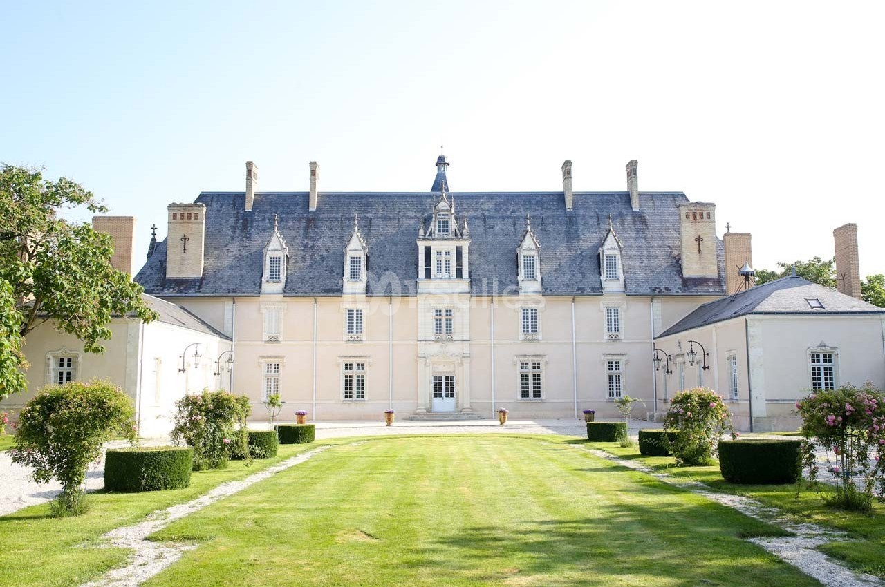 Façade d'un château avec toit en ardoise, entouré de jardins bien entretenus et d'arbustes taillés.