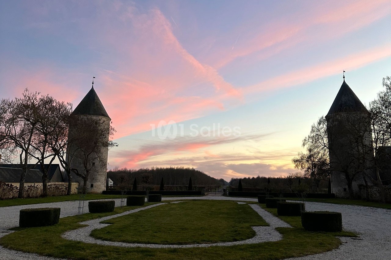 Deux tours en pierre entourant une allée bordée d'arbres, sous un ciel de coucher de soleil aux teintes rosées.