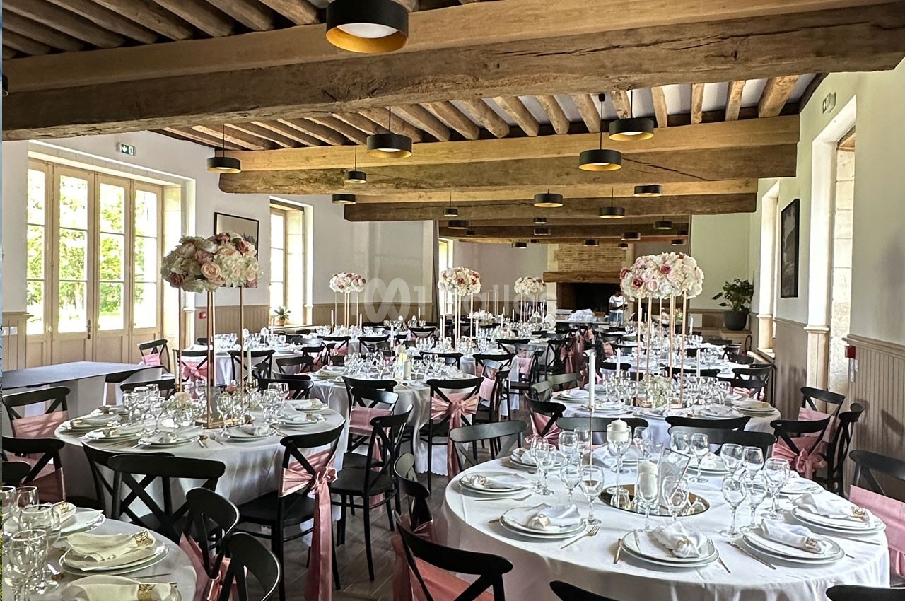 Salle de réception décorée pour un événement, avec des tables rondes dressées et des centres de table fleuris.