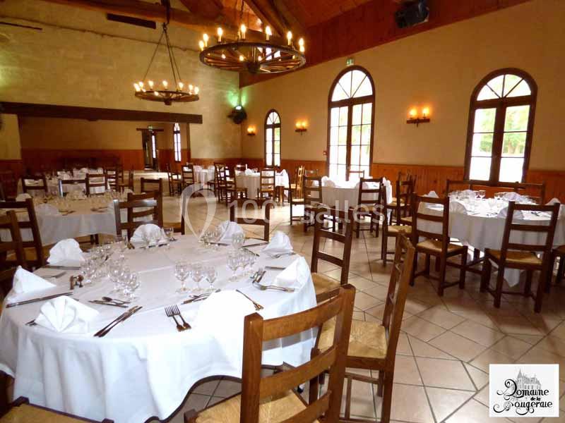 Salle de réception avec tables rondes dressées, nappes blanches, chaises en bois et lumière naturelle entrant par les…