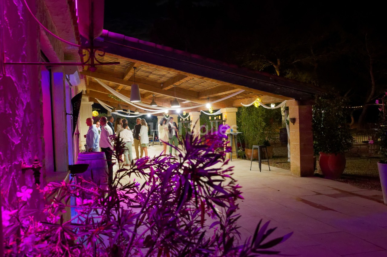 Terrasse éclairée en soirée avec des invités discutant sous un auvent décoré, entourée de végétation.
