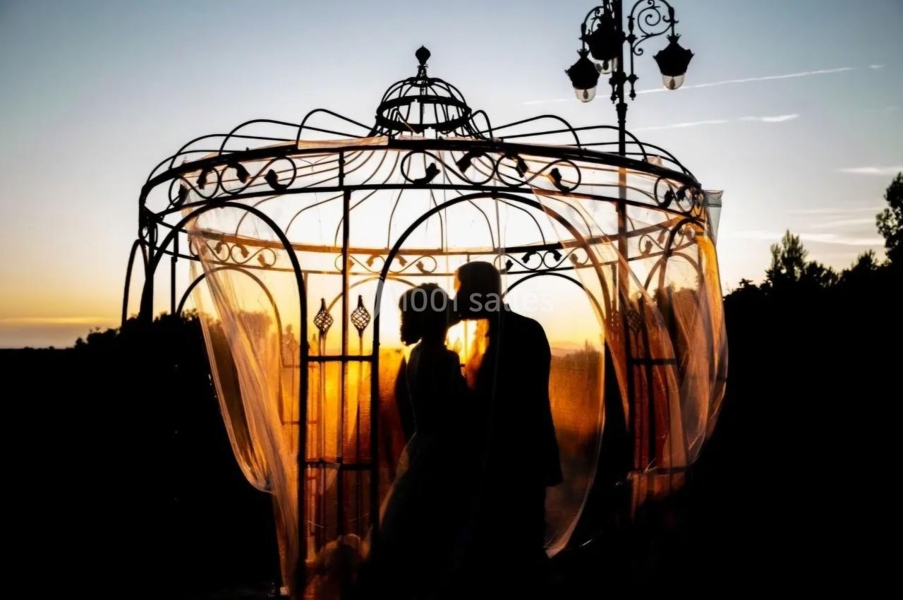 Un couple se tient sous une structure en fer forgé au coucher du soleil, entouré de voilages translucides.