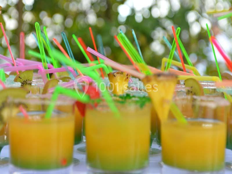 Verres de cocktails colorés garnis de fruits et pailles multicolores, disposés sur une table en extérieur.
