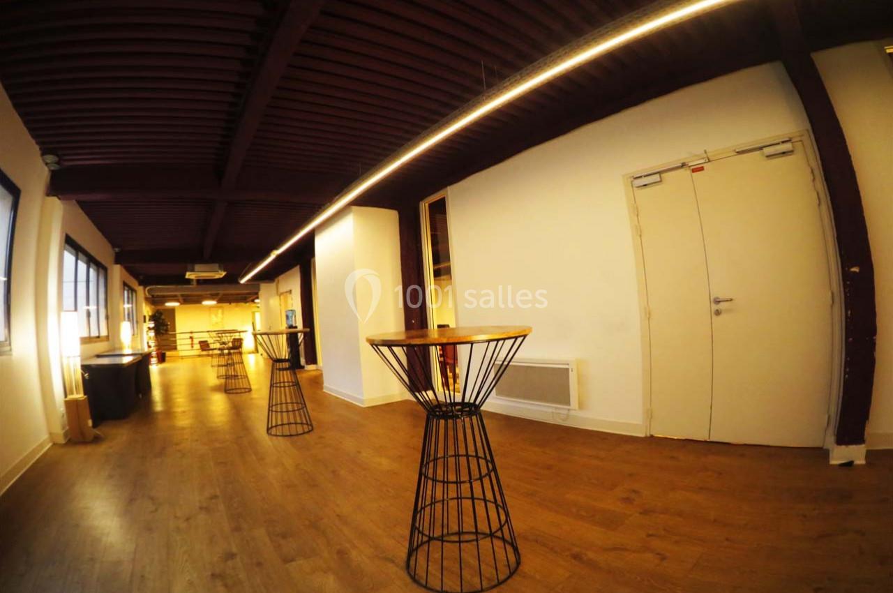 Salle avec parquet, tables hautes en métal et bois, plafond à poutres apparentes, éclairage chaleureux.