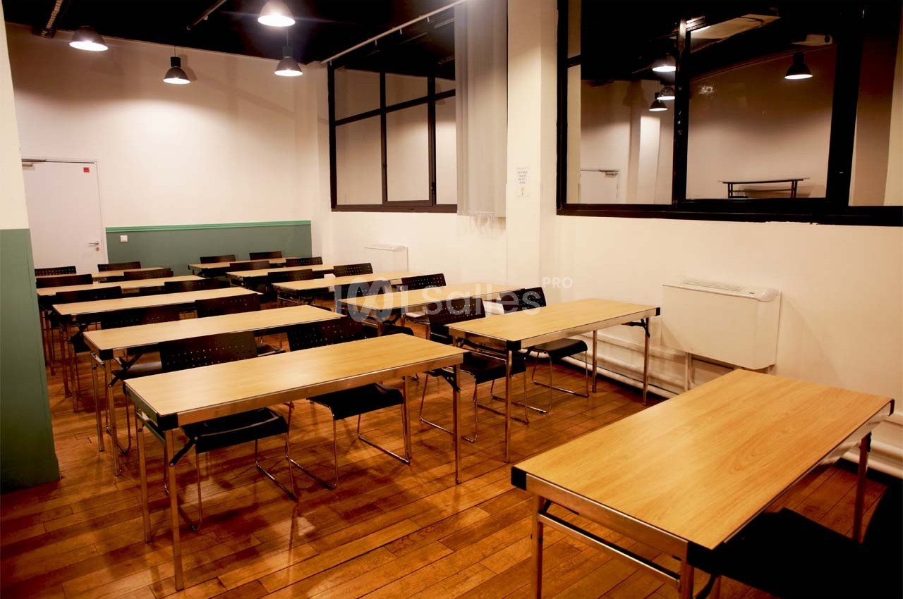 Salle de classe vide avec des tables en bois, des chaises noires et un éclairage suspendu.