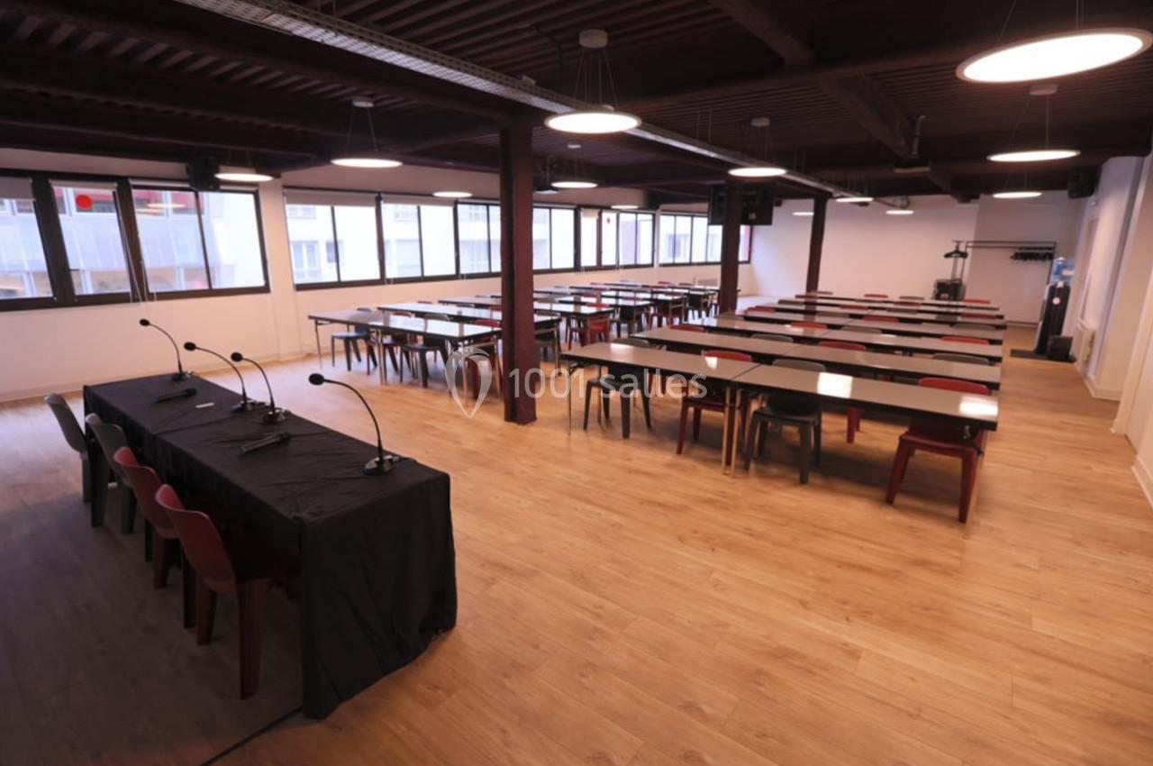 Salle de conférence vide avec tables, chaises, micros et éclairage suspendu, parquet au sol et grandes fenêtres.