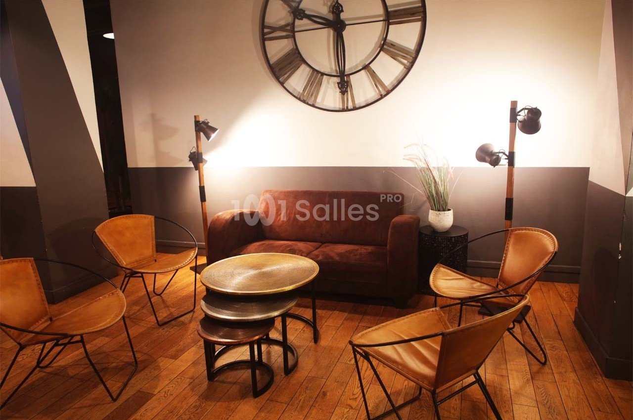 Espace salon avec canapé en velours marron, chaises en cuir, tables rondes et grande horloge murale.