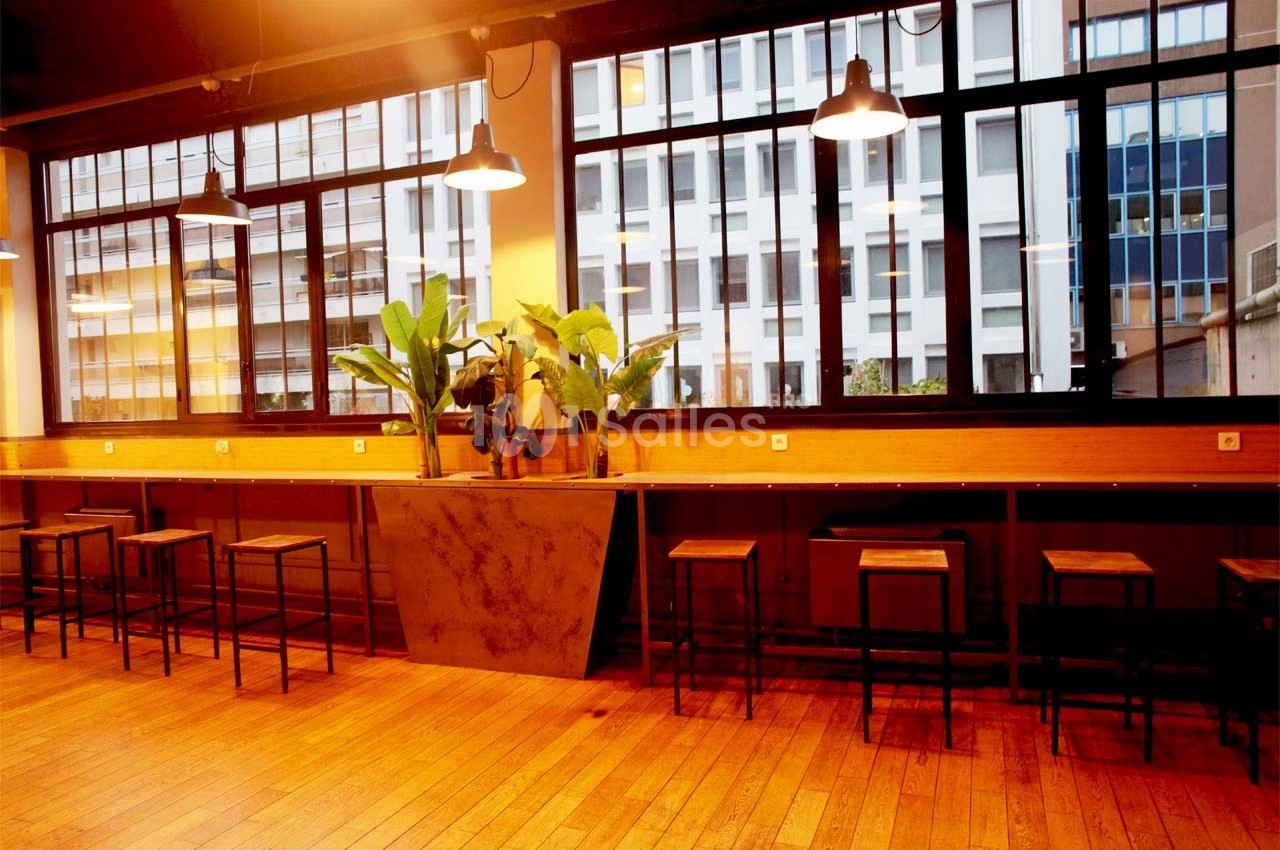 Salle lumineuse avec de grandes fenêtres, des tabourets en bois et métal alignés le long d'un comptoir avec des plantes.