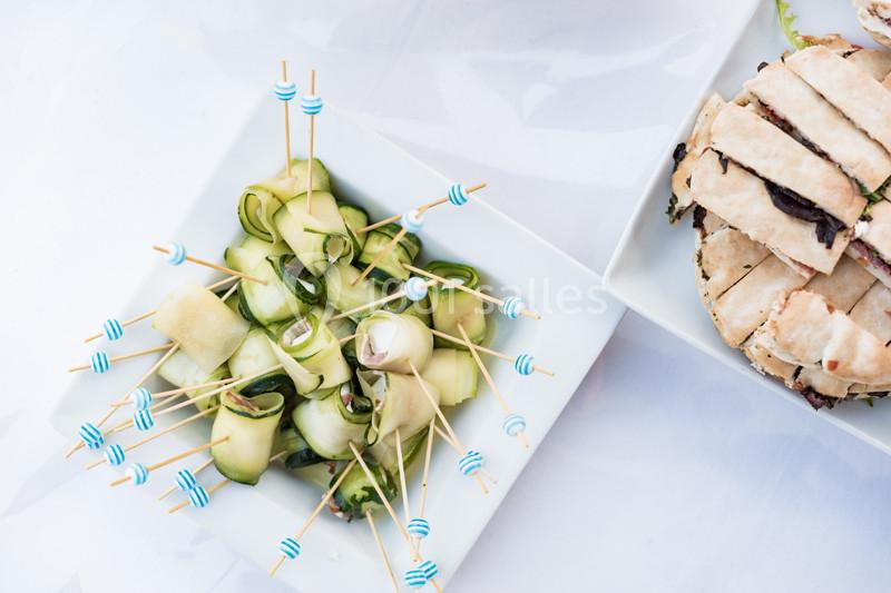 Assortiment d'amuse-bouches avec roulés de courgettes sur piques et morceaux de pain garnis sur assiettes blanches.