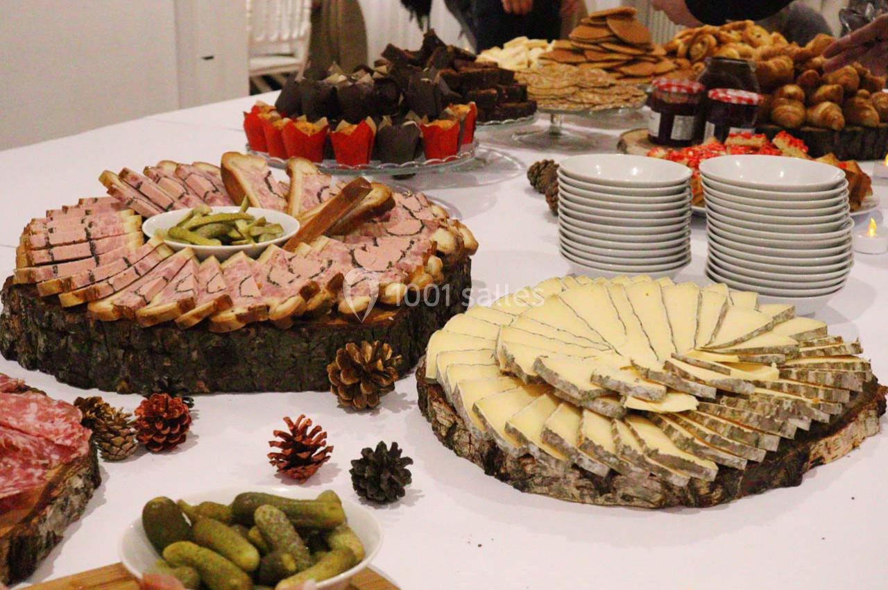 Plateau de charcuterie, fromage, cornichons et pains sur une table, entourés de desserts et vaisselle.