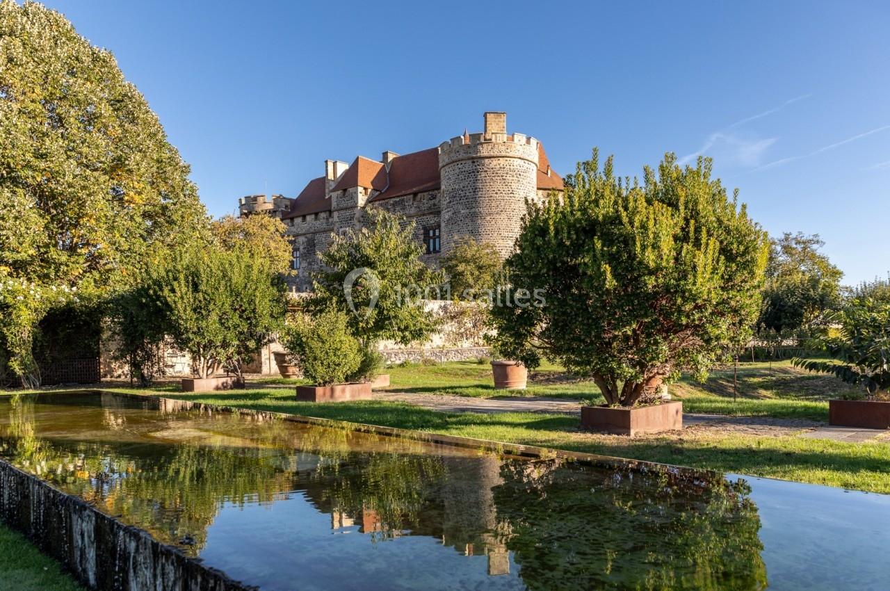 Vue d'un château en pierre entouré de jardins arborés, avec un bassin reflétant le bâtiment sous un ciel dégagé.