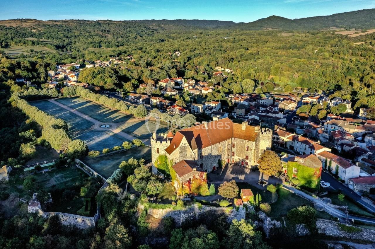 Vue aérienne d'un château médiéval entouré de jardins, avec un village et des collines verdoyantes en arrière-plan.