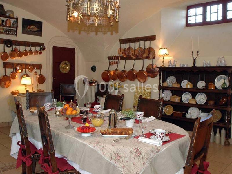 Salle à manger rustique avec une table dressée, des chaises en bois, des plats variés et des ustensiles en cuivre accrochés…