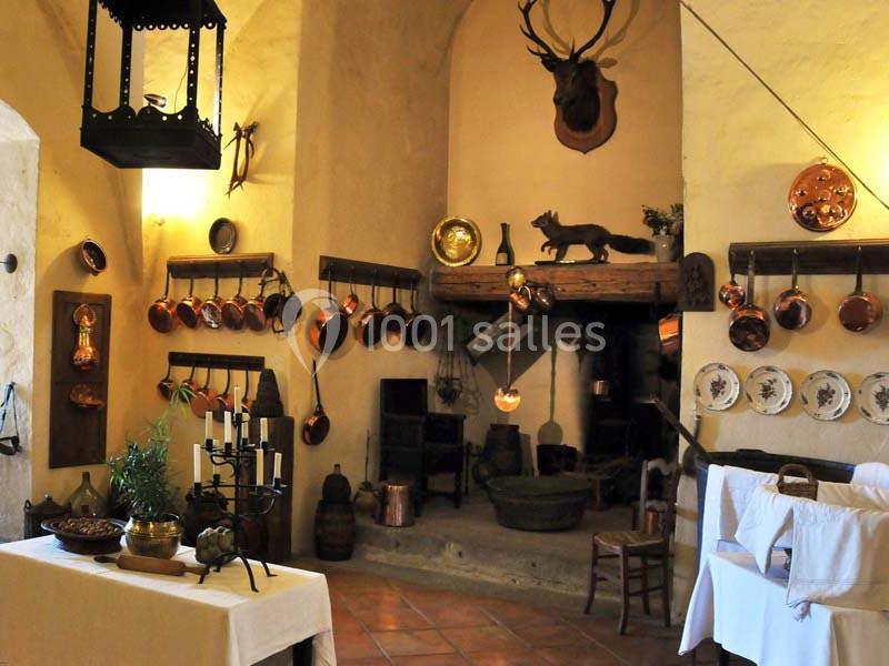 Cuisine ancienne avec murs en pierre, ustensiles en cuivre accrochés, cheminée rustique et table dressée.