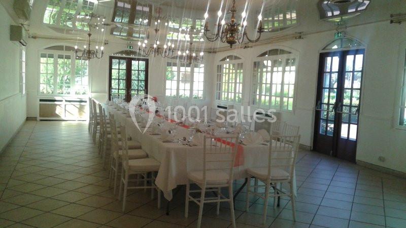 Salle lumineuse avec une grande table dressée pour un repas, entourée de chaises blanches et éclairée par des lustres.