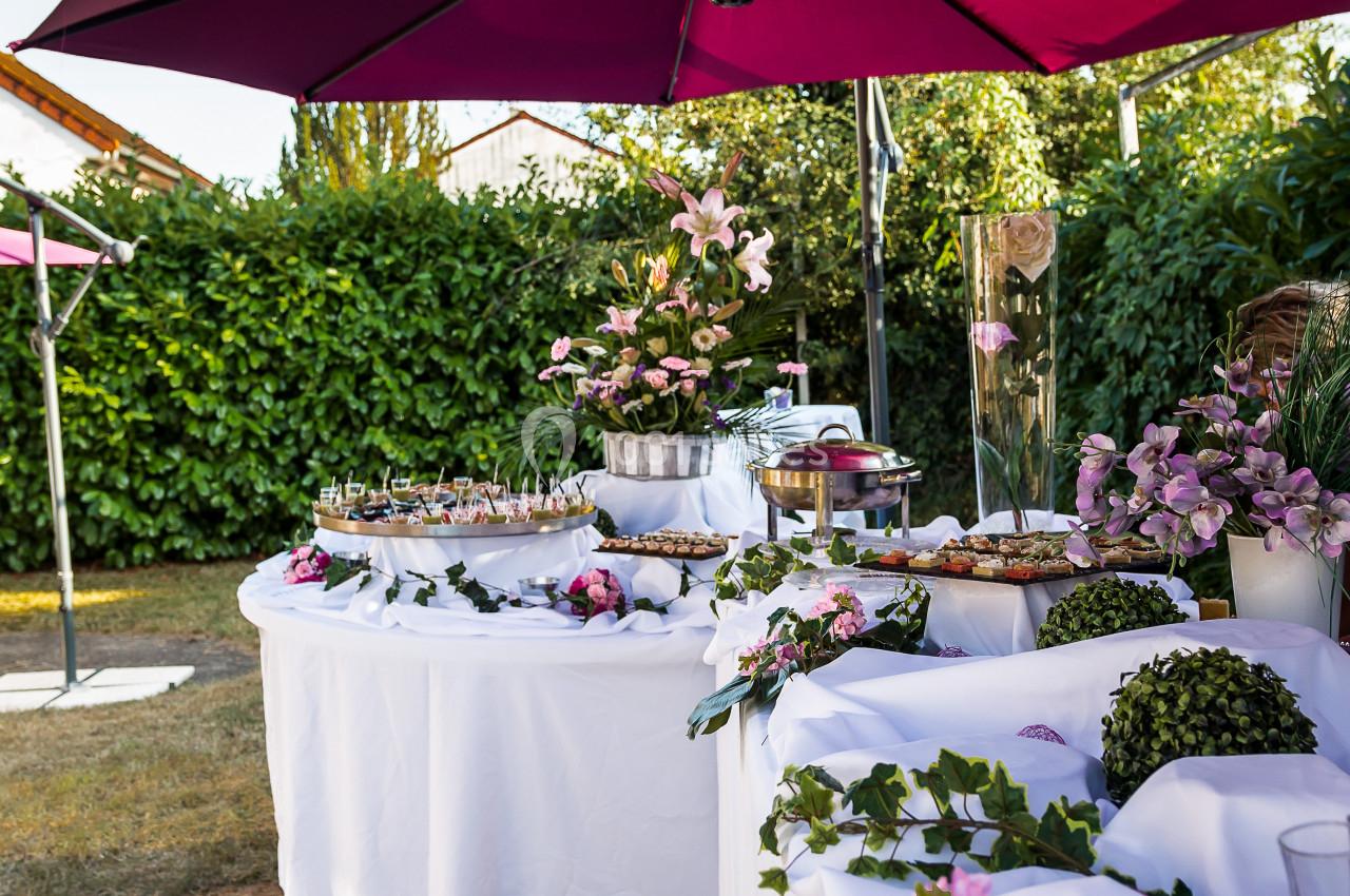 Buffet en plein air avec nappes blanches, fleurs roses et parasols violets, entouré de verdure.