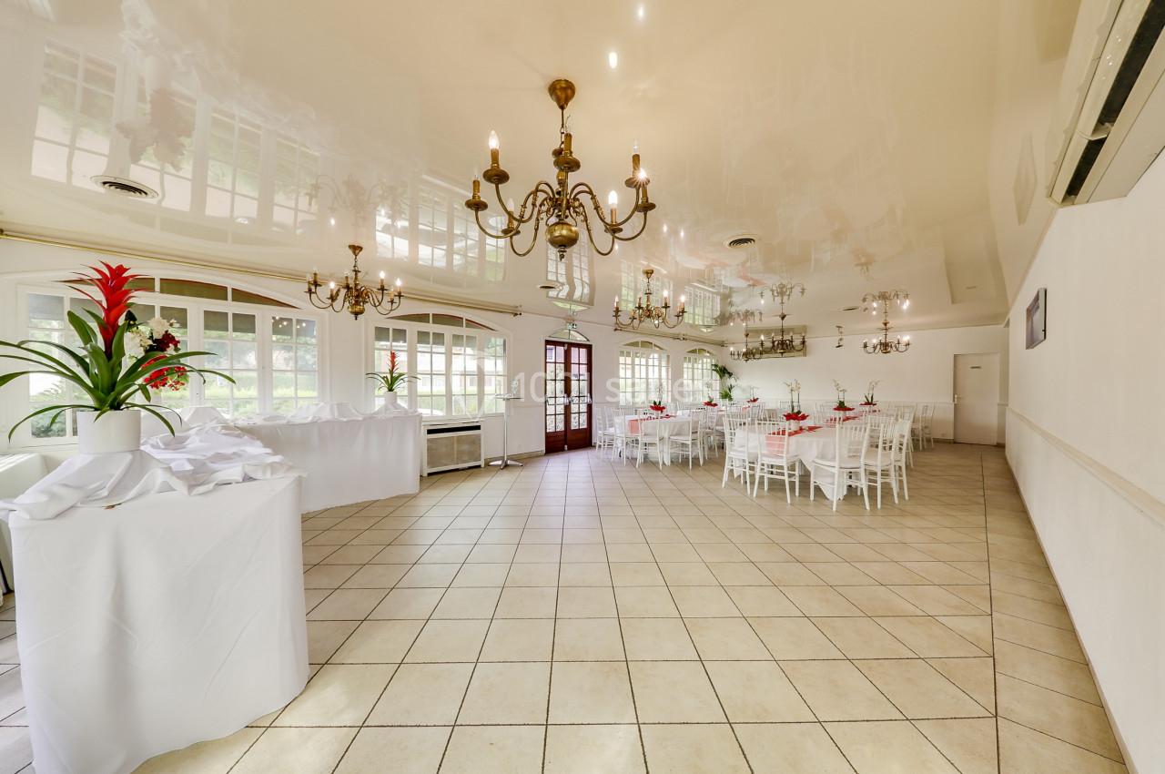 Salle de réception lumineuse avec tables dressées, chaises blanches, lustres et décorations florales rouges.