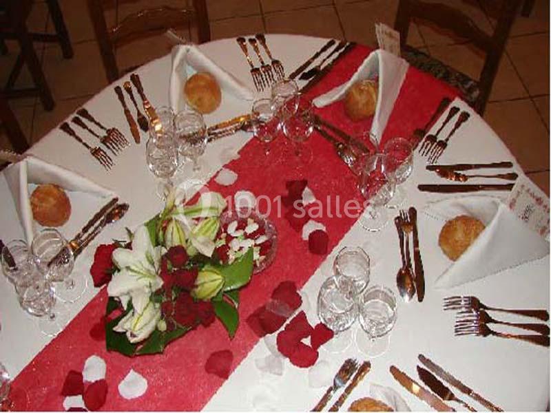Table ronde dressée avec nappes blanches, chemin de table rouge, fleurs, pétales et couverts disposés pour un repas.
