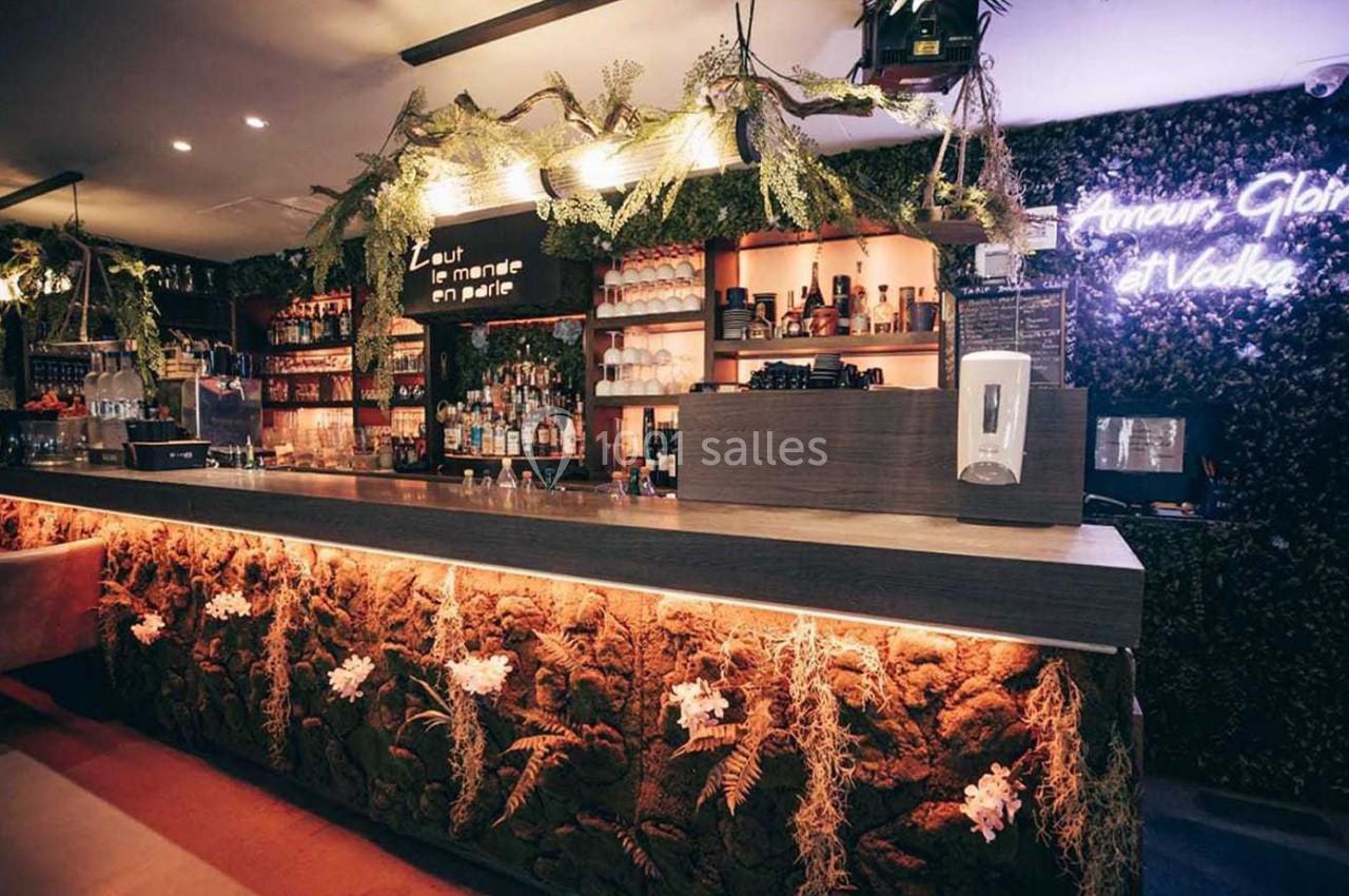 Bar moderne avec comptoir éclairé, décor végétal, étagères de bouteilles et néon lumineux au mur.