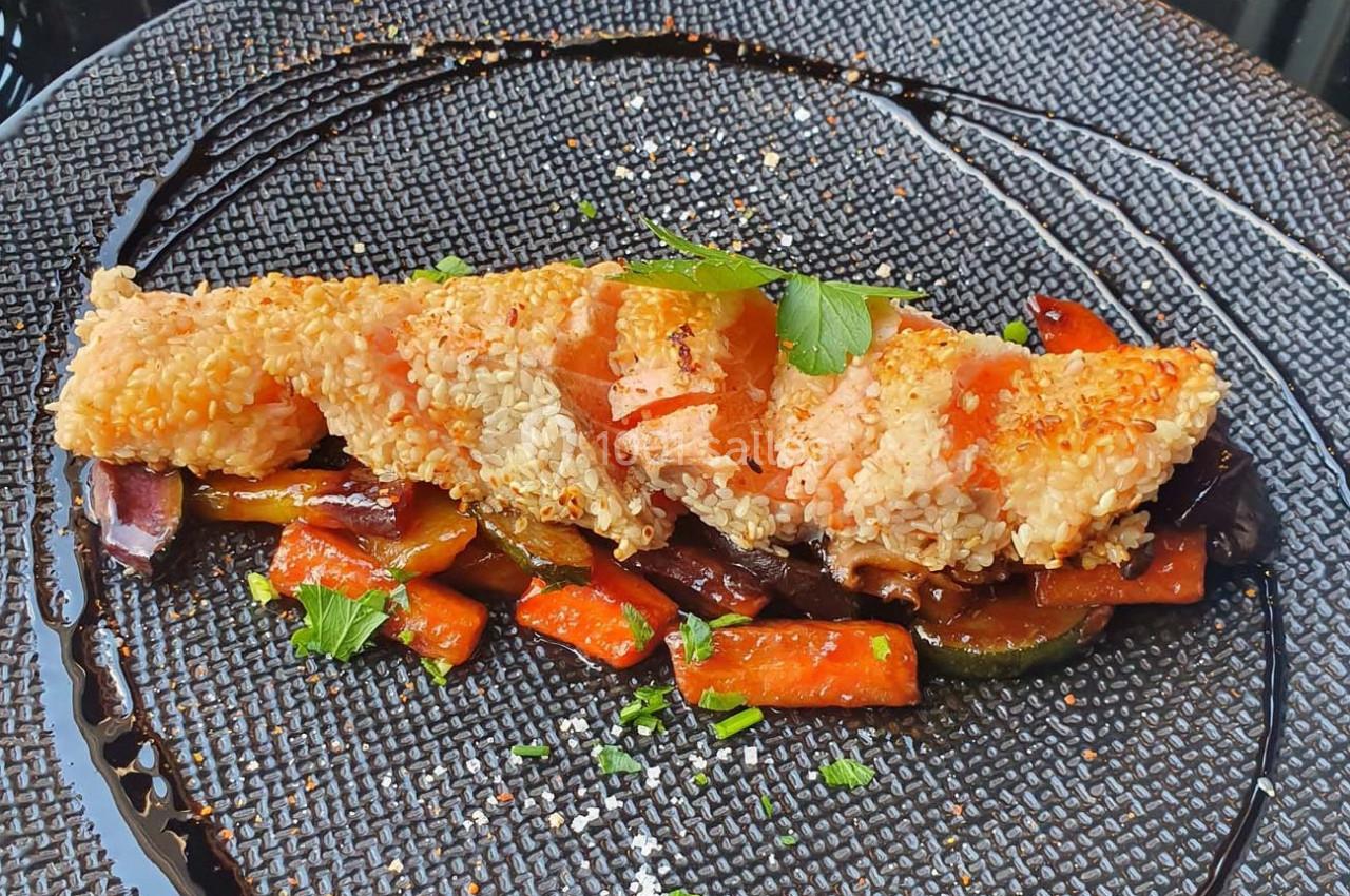 Filet de poisson pané au sésame servi sur un lit de légumes grillés, décoré de persil et d'un filet de sauce.