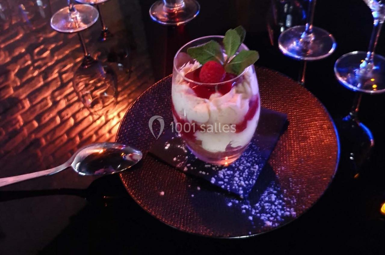 Verre de dessert composé de crème, framboises et feuilles de menthe, posé sur une assiette noire avec une cuillère.