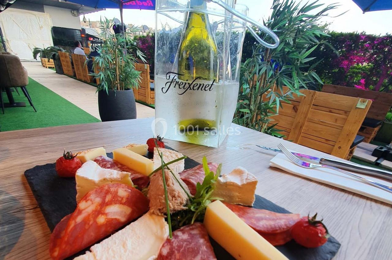 Planche de charcuterie et fromages sur une table en terrasse, avec une bouteille de vin blanc dans un seau à glace.