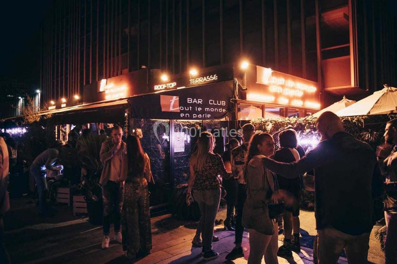 Groupe de personnes discutant et se détendant devant un bar éclairé en soirée, avec une ambiance animée.