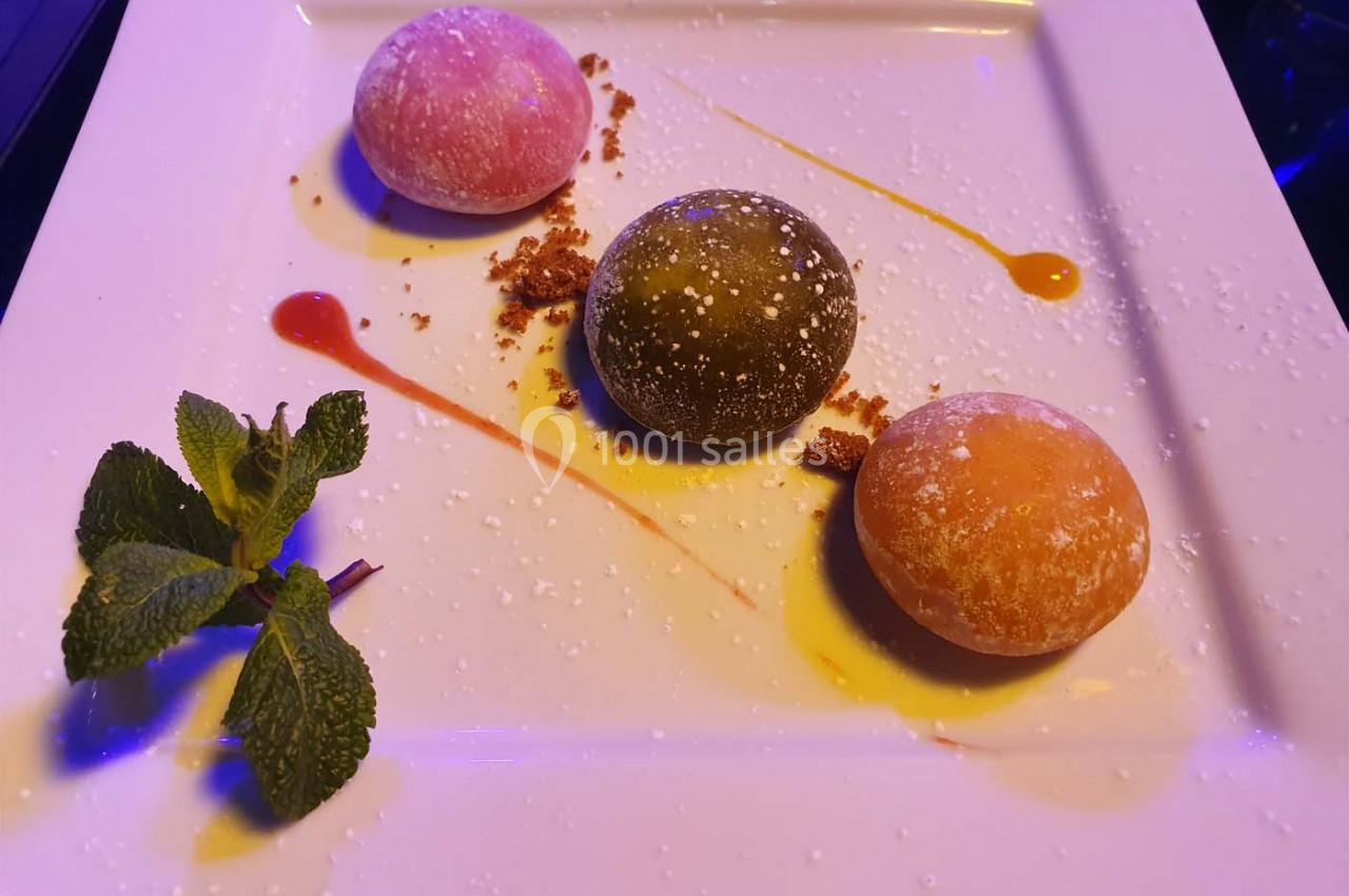 Assiette contenant trois mochis colorés, décorée de coulis, miettes de biscuit et une branche de menthe.