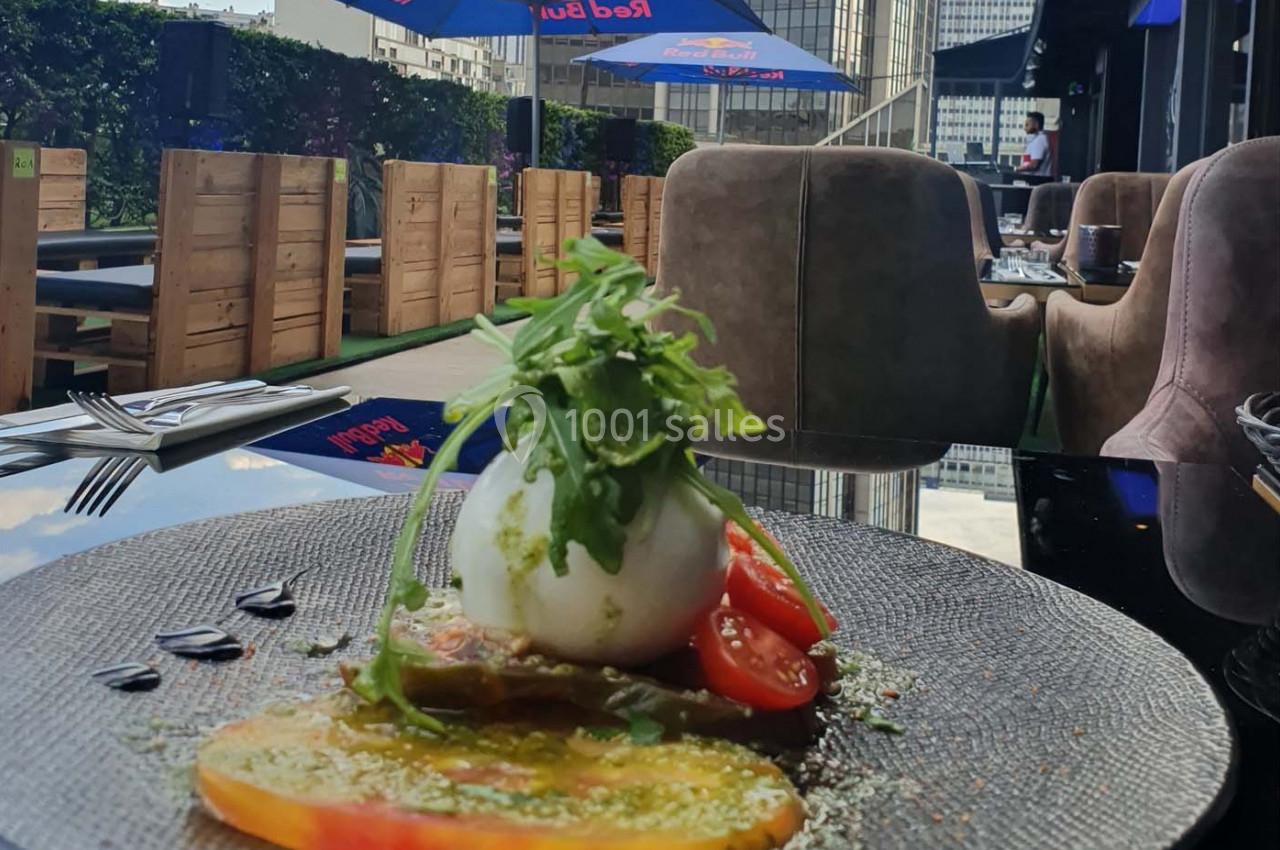 Assiette de burrata avec tomates colorées, roquette et filet de pesto, servie en terrasse avec vue sur des immeubles.