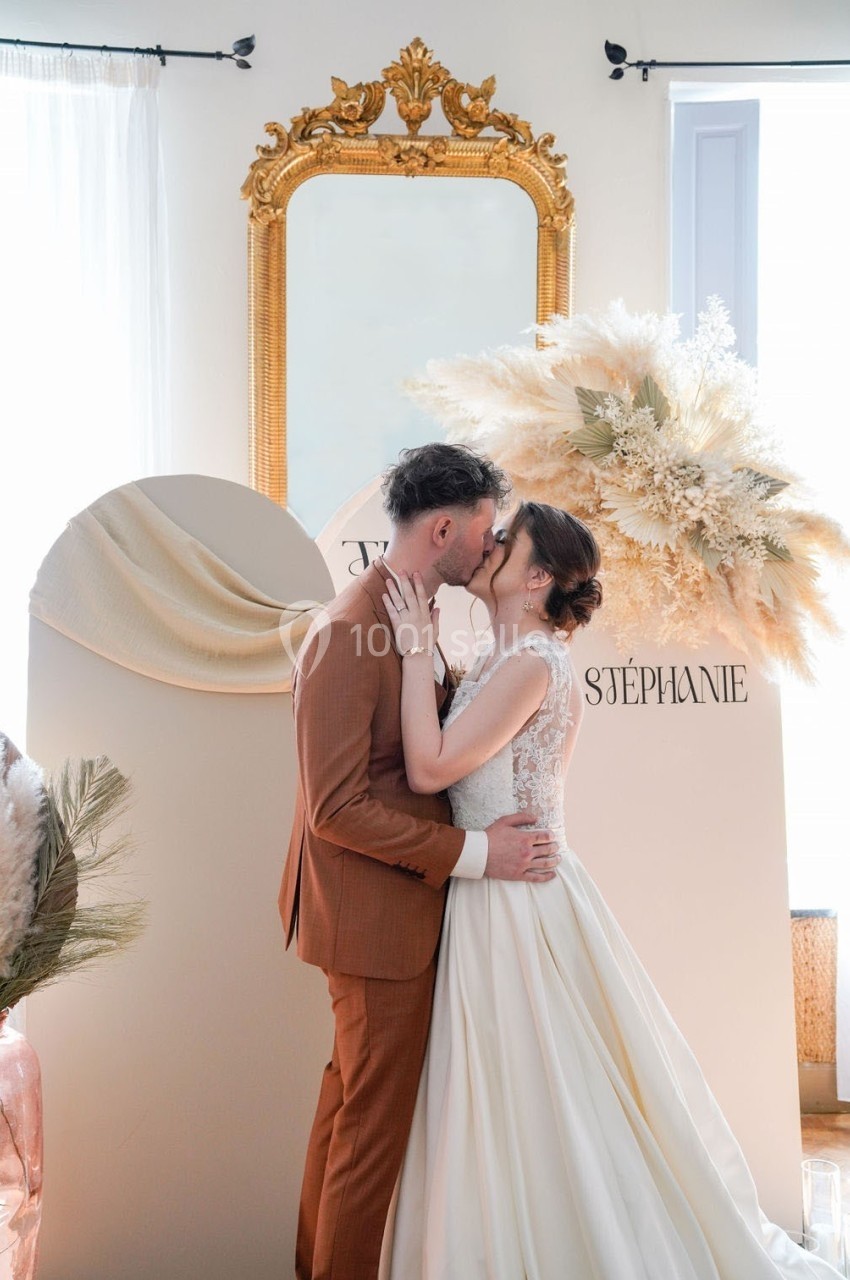 Un couple de mariés s'embrasse devant une décoration élégante avec un miroir doré et des fleurs séchées.
