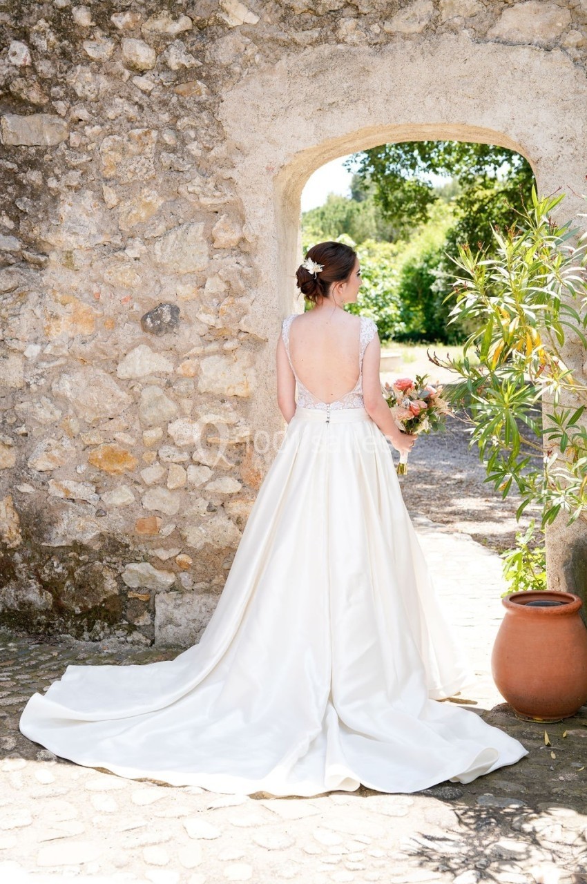 Une femme en robe de mariée blanche, vue de dos, debout sous une arche en pierre, tenant un bouquet de fleurs.