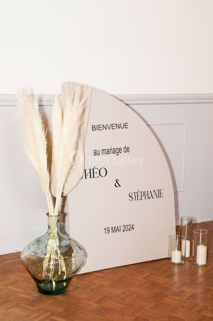 Panneau de bienvenue pour un mariage, posé au sol près d'un vase avec des herbes séchées et des bougies.