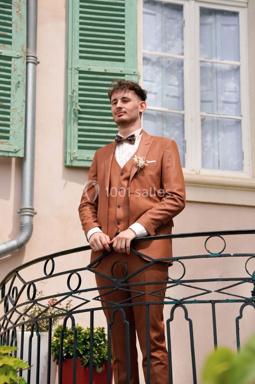 Homme en costume marron se tenant sur un balcon en fer forgé devant une façade beige avec volets verts.