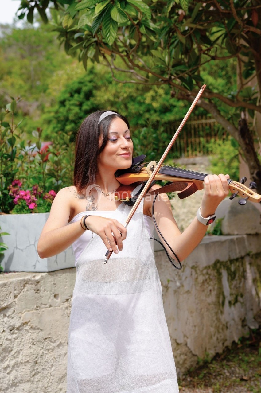 Une femme joue du violon en extérieur, entourée de verdure et de fleurs, près d'un muret en pierre.