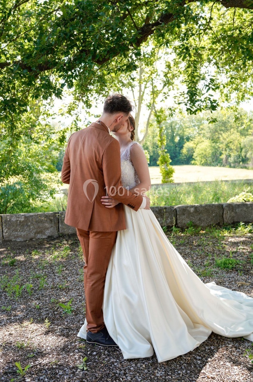 Un couple en tenue de mariage s'enlace sous un arbre dans un cadre naturel et ensoleillé.